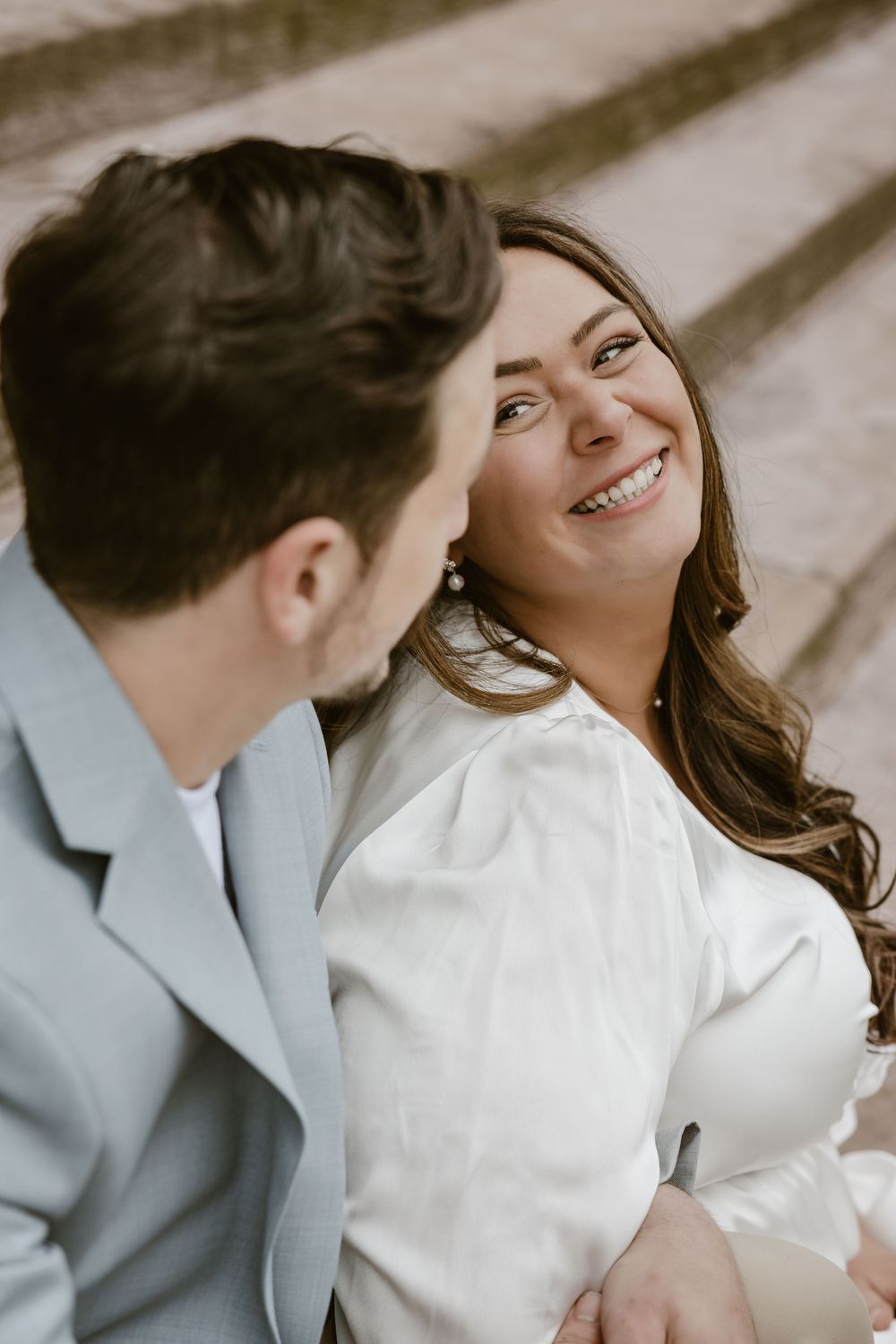 Romantic portrait of couple sitting on steps with person in gray suit gazing at smiling partner in white.