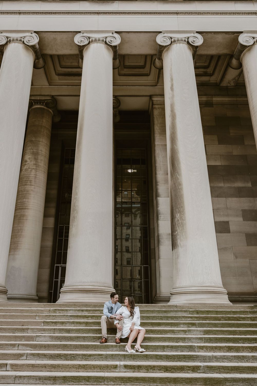 Two people sit on grand stone steps beneath towering neoclassical columns of a historic building.