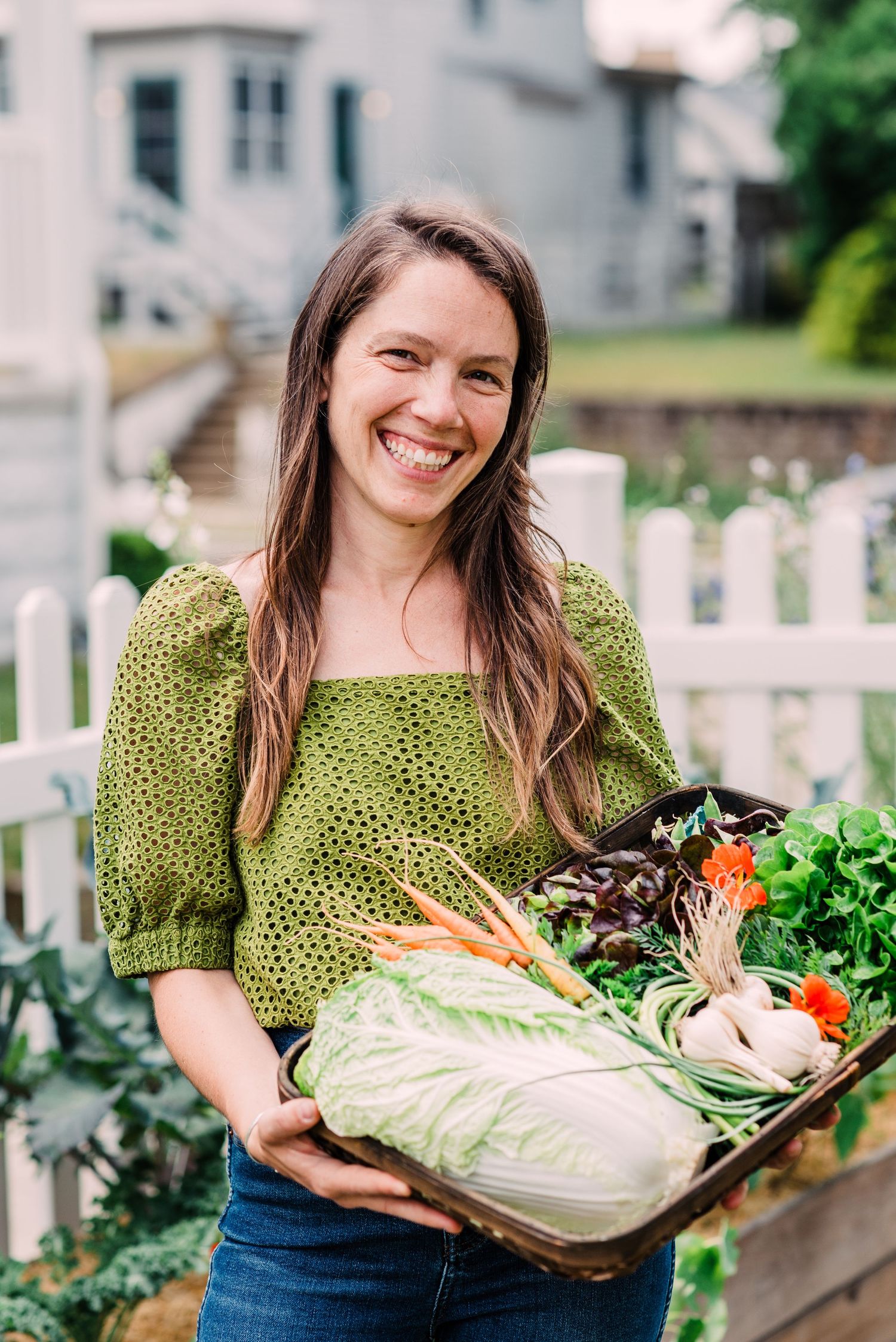 In a green sweater, a person displays a basket filled with fresh garden vegetables against a white picket fence.