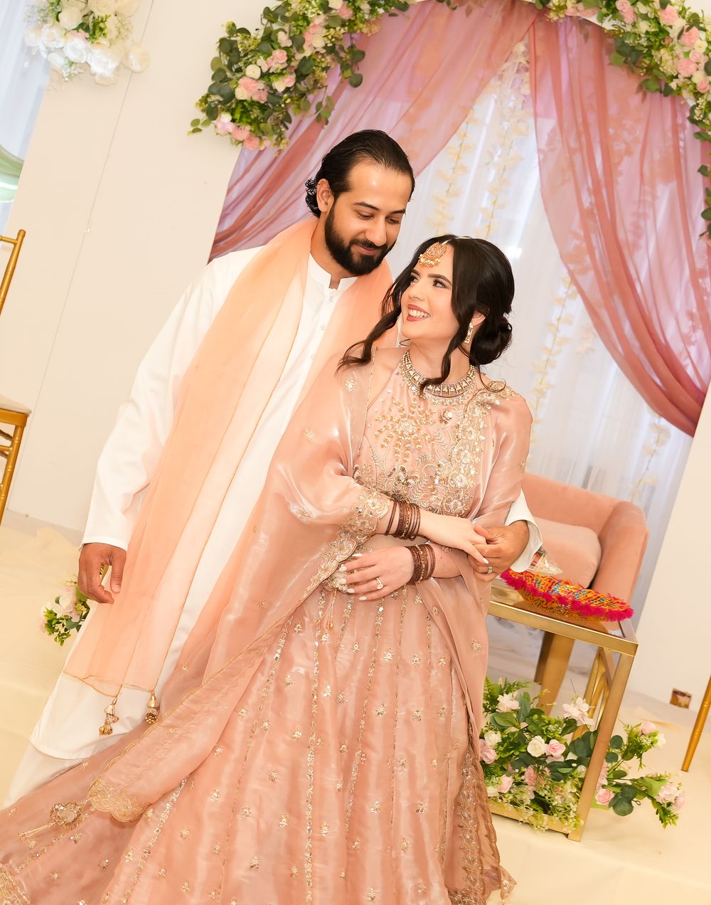 Couple in matching blush pink South Asian wedding attire standing under a floral arch at their engagement ceremony.