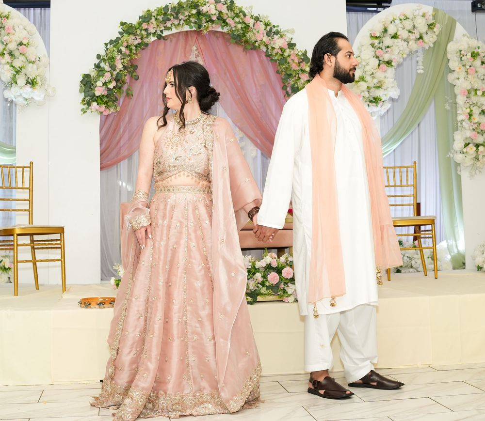 South Asian bride and groom in pink lehenga and kurta holding hands at floral wedding arch.