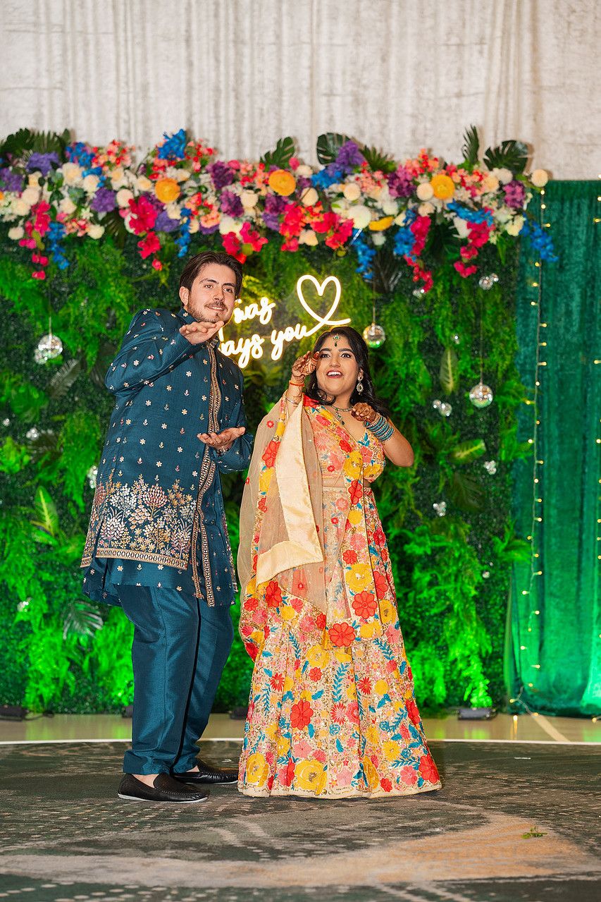 Couple in vibrant Indian attire posing before a floral backdrop with neon heart sign at a festive celebration.