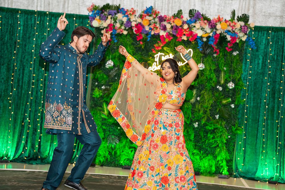 Couple dancing joyfully at an Indian wedding celebration with colorful floral backdrop and festive traditional attire.