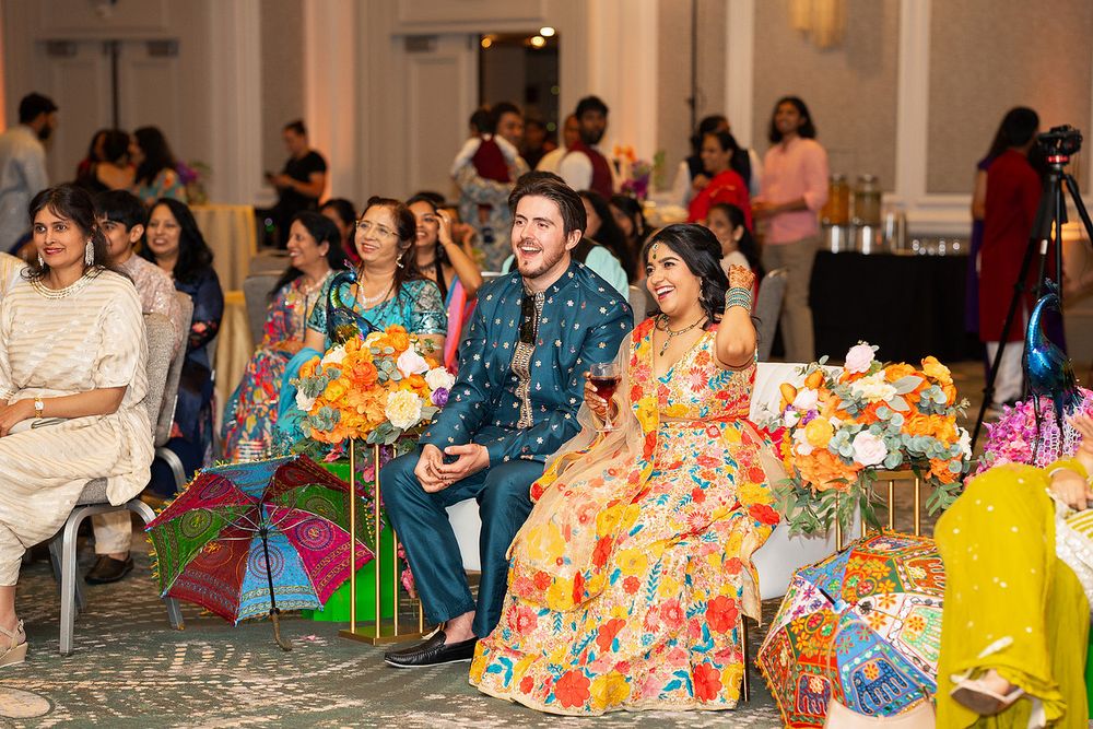 Couple in vibrant traditional attire laughing at colorful South Asian wedding celebration with guests and floral decor.