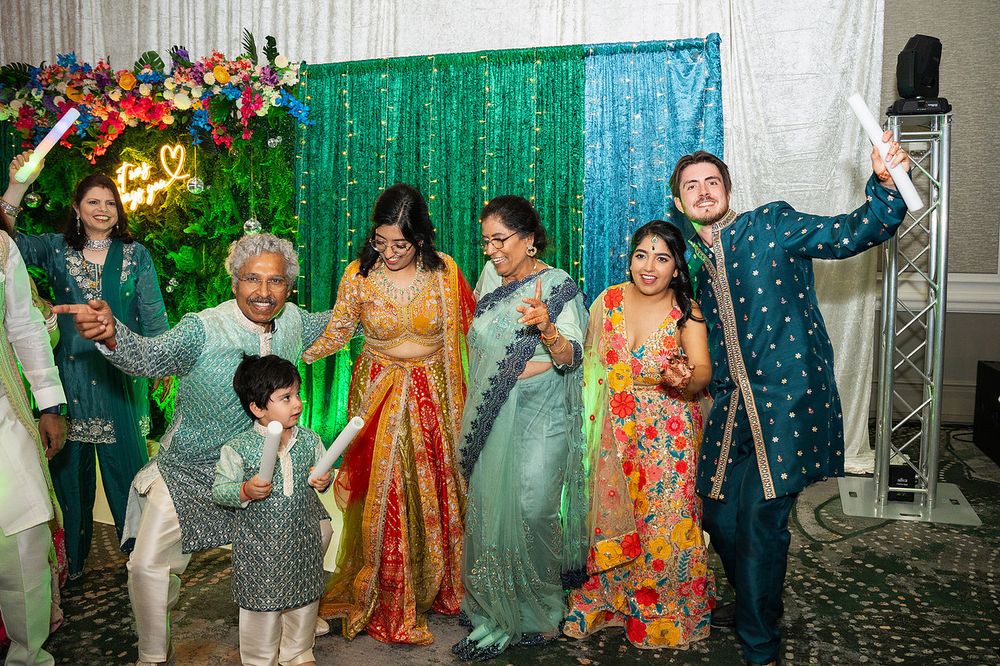 Joyful Indian wedding celebration with guests in colorful traditional attire posing together against a green sequin backdrop.