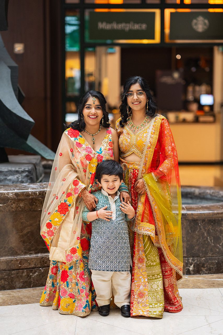Two women in colorful Indian sarees pose with a young boy in traditional attire near a Starbucks Marketplace.