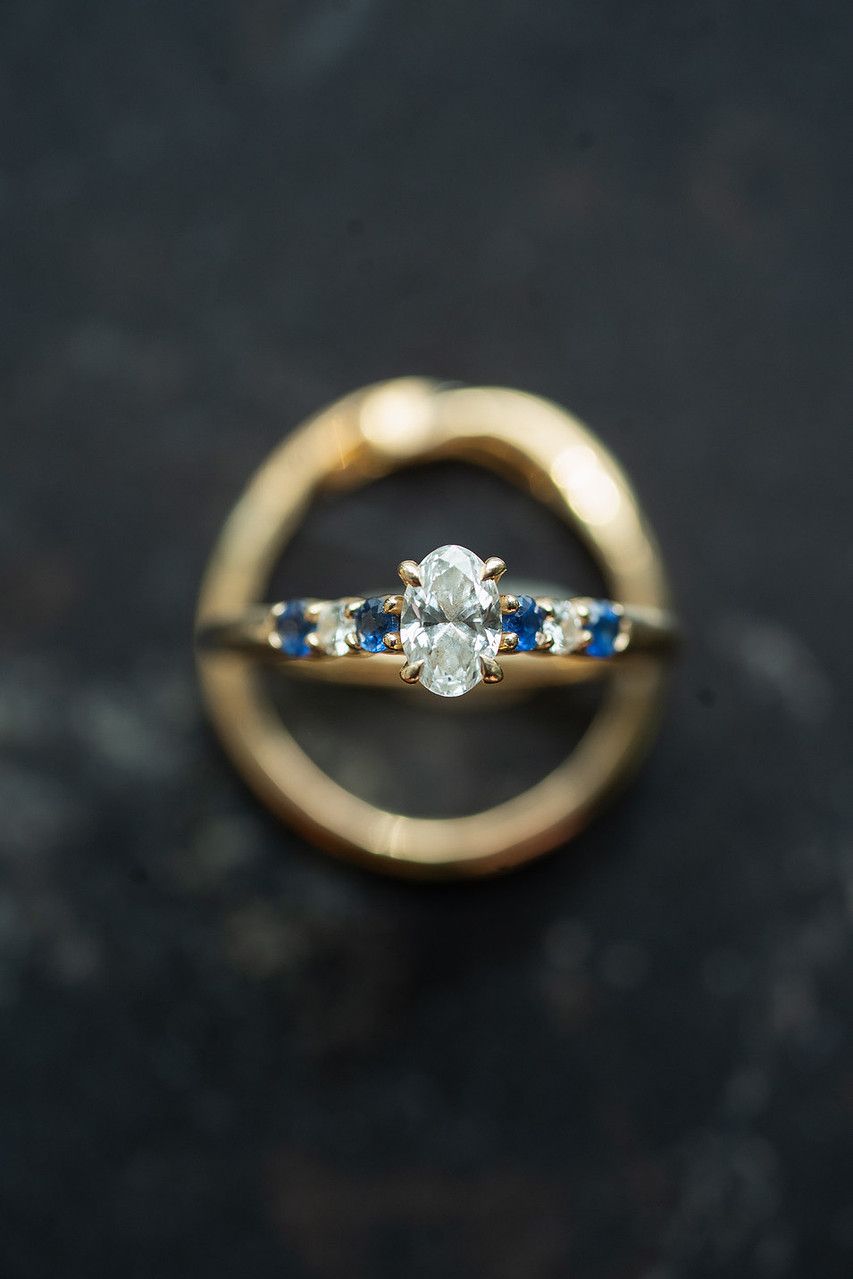 Gold engagement ring with oval diamond center stone and blue sapphire accents, nestled inside a plain gold band on dark surface.