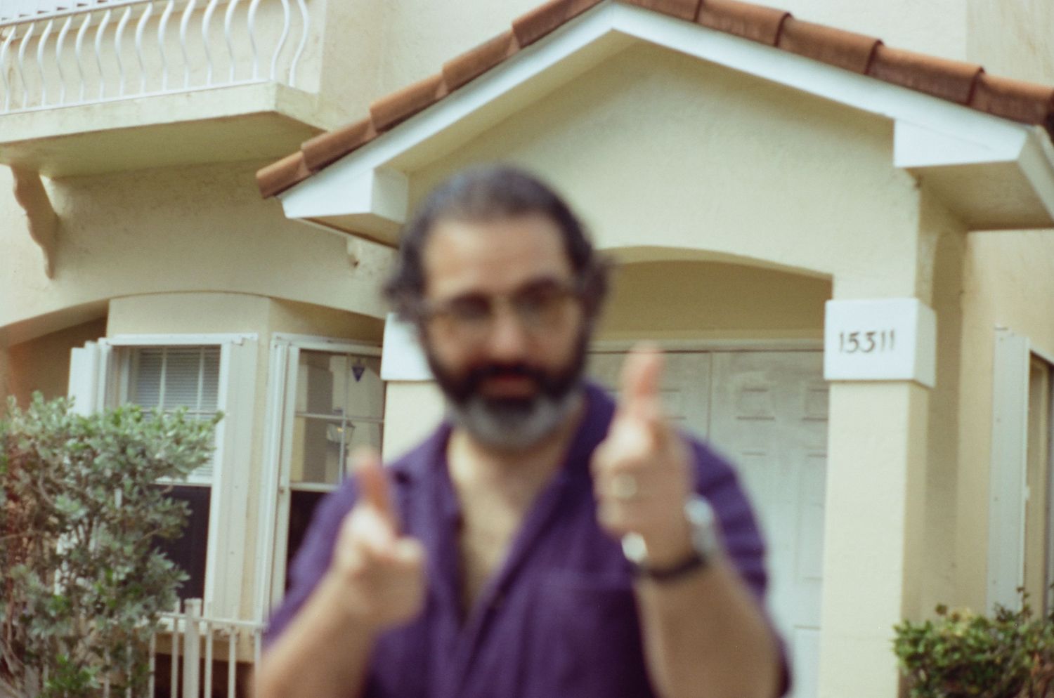 A person wearing a purple shirt gives two thumbs up while standing in front of a beige house.