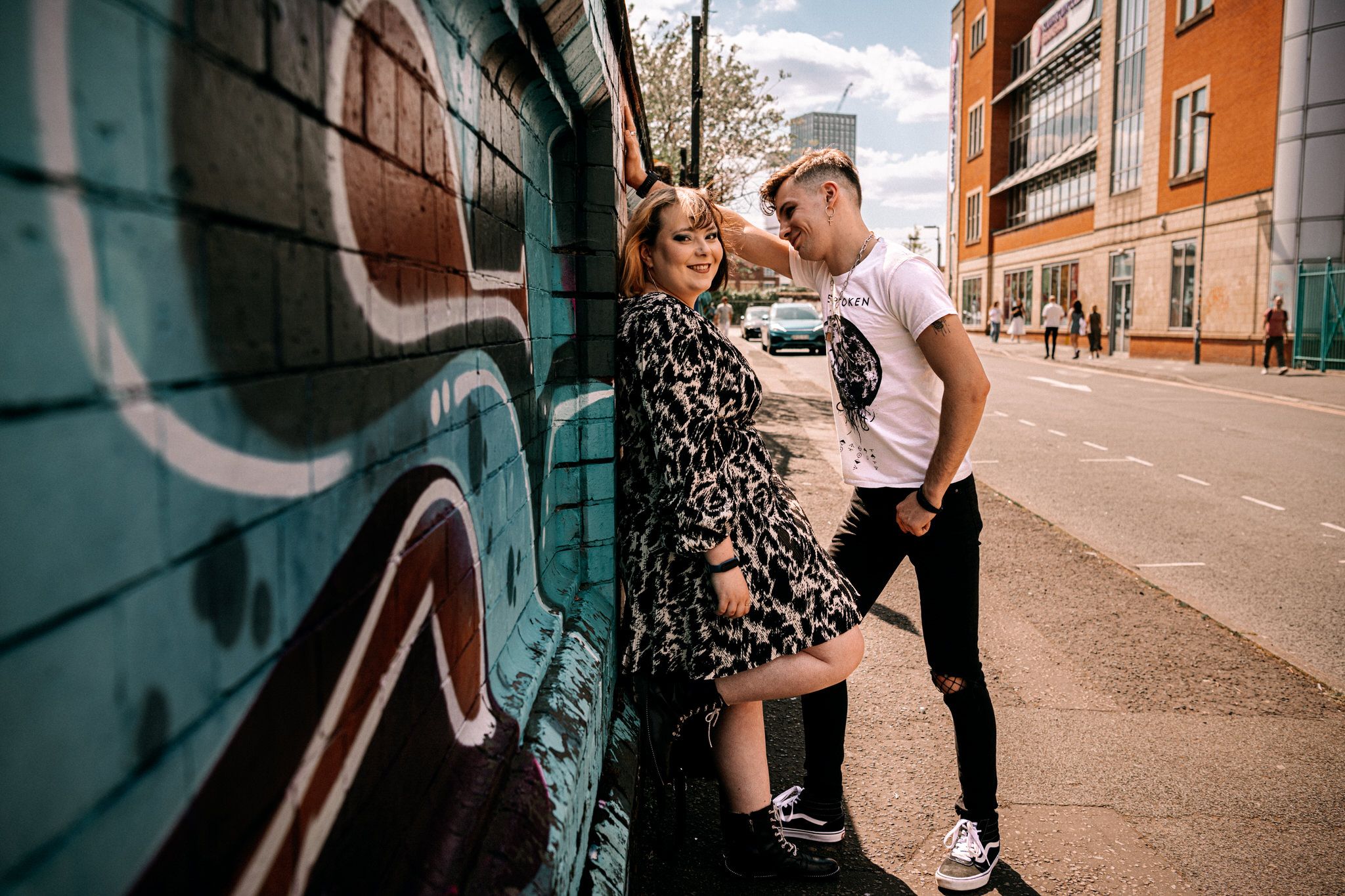 A couple shares an intimate moment against a graffiti wall on an urban street with orange buildings in the background.