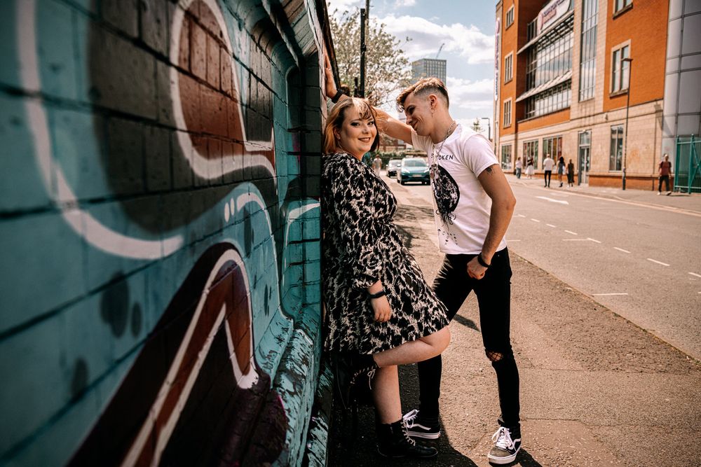 A couple shares an intimate moment against a graffiti wall on an urban street with orange buildings in the background.