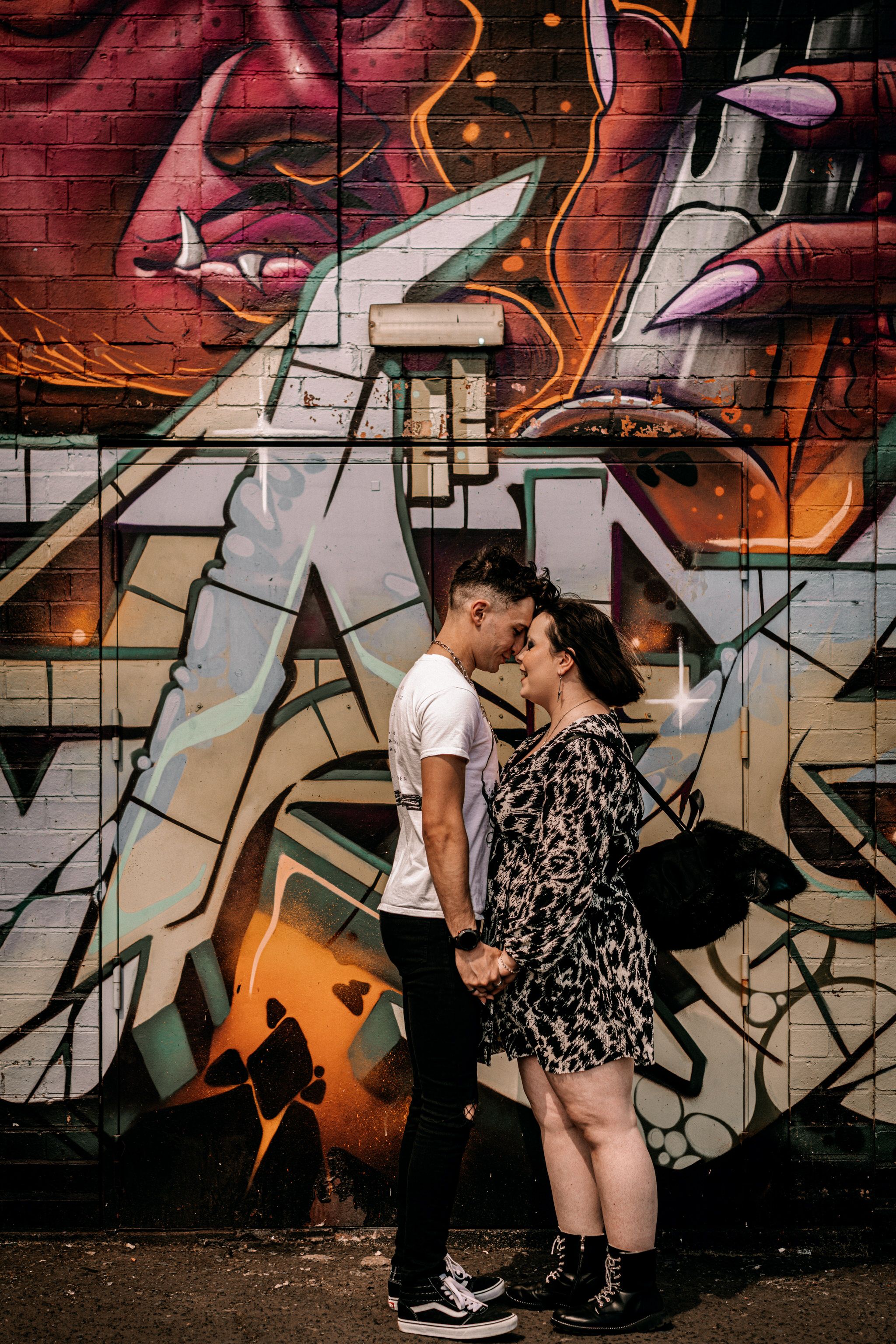 A romantic couple shares an intimate moment against a vibrant graffiti wall with orange and white street art designs.