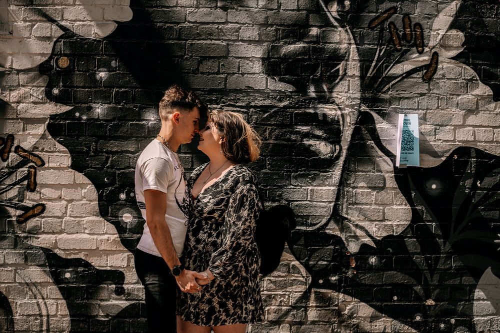A romantic couple embraces against an urban brick wall featuring dramatic black and white street art murals.