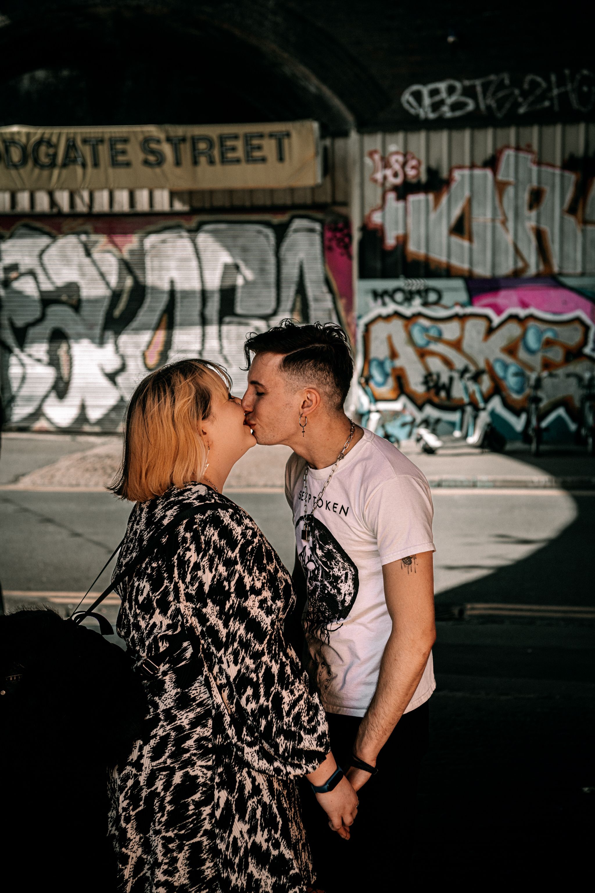 A couple shares a romantic kiss against a graffiti-covered wall in an urban street setting.
