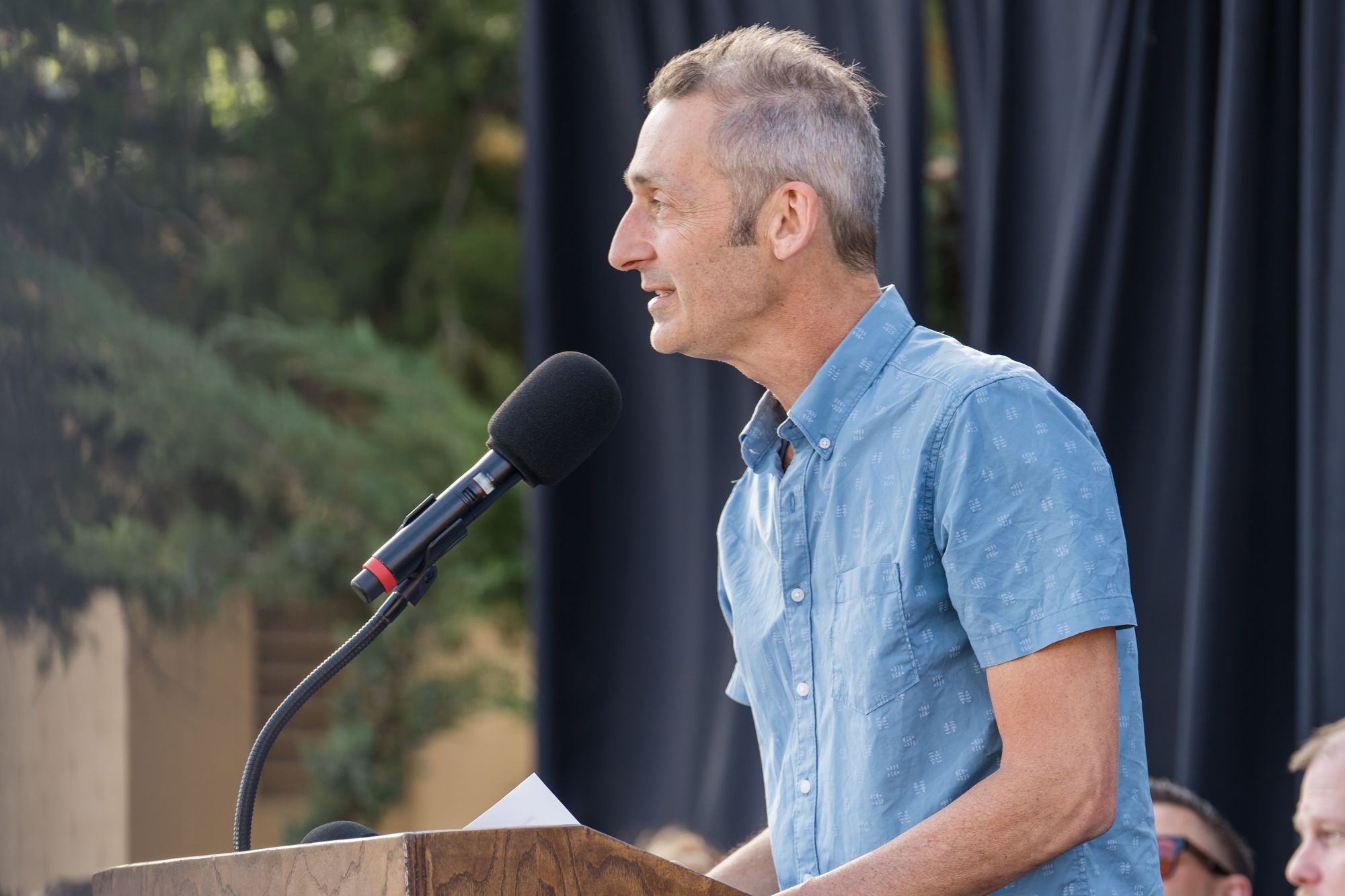 Someone speaks from a wooden podium into a microphone while wearing a light blue short-sleeved shirt on an outdoor stage.