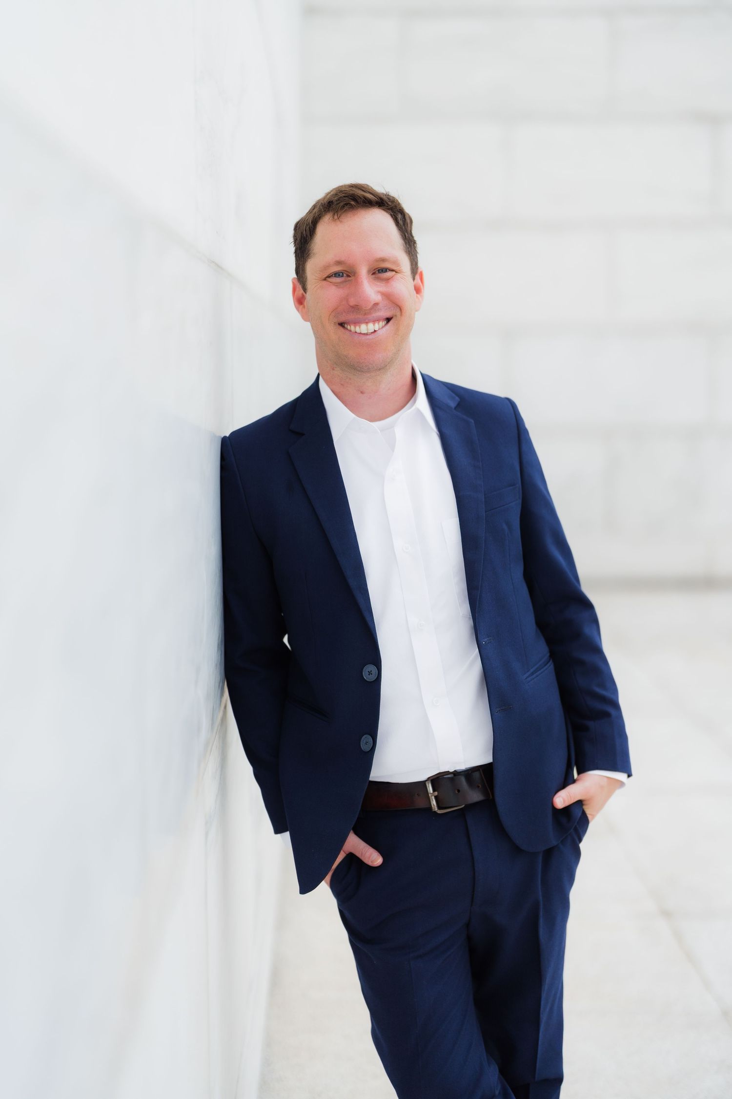 Professional portrait of someone in a navy blue suit with white shirt standing against a white wall in casual pose.