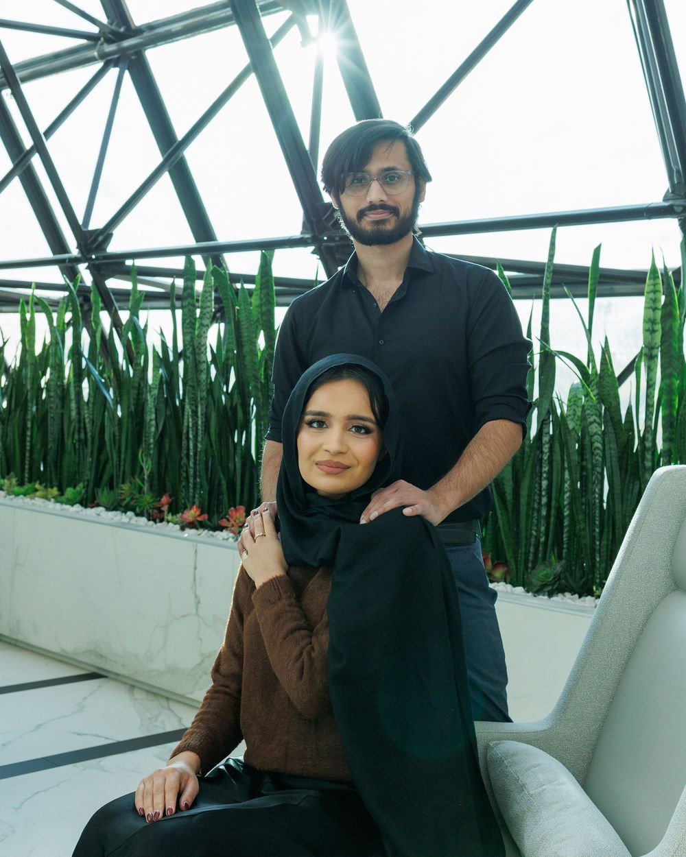 A couple poses together in a greenhouse setting with tall green plants and industrial architecture visible in background.