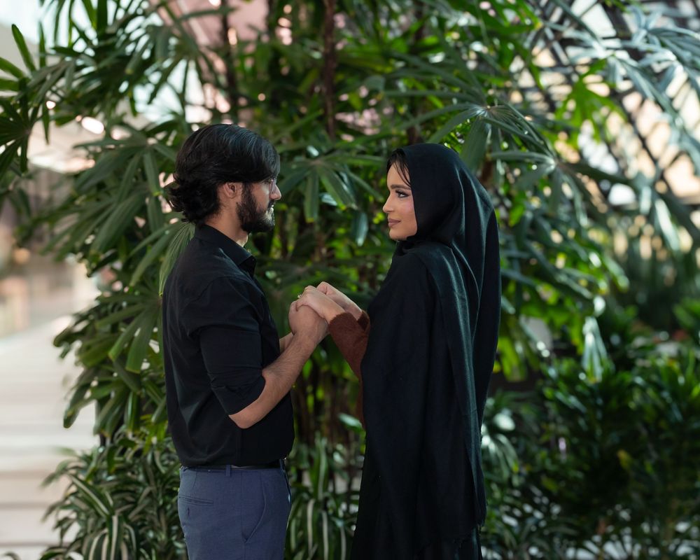 Two people in dark clothing hold hands while standing near lush green plants in an intimate scene.