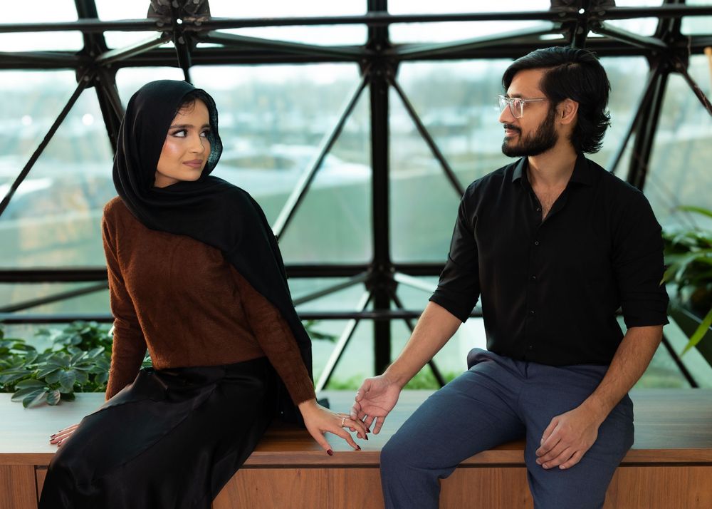 Two people sit together in a modern glass atrium with city views as one wears a brown hijab and the other a black shirt.