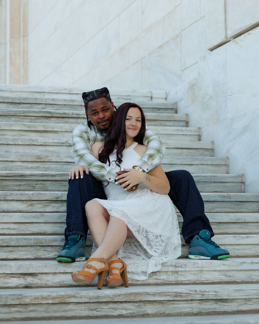 A couple sits together on white marble steps wearing jeans and boots in a romantic engagement photo series.