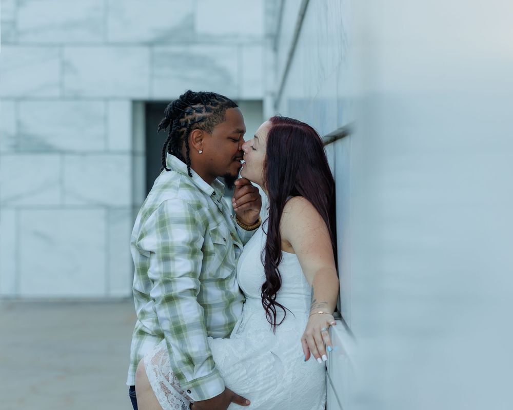 Couple in romantic pose against white wall, with one wearing plaid button-up and other in white dress.
