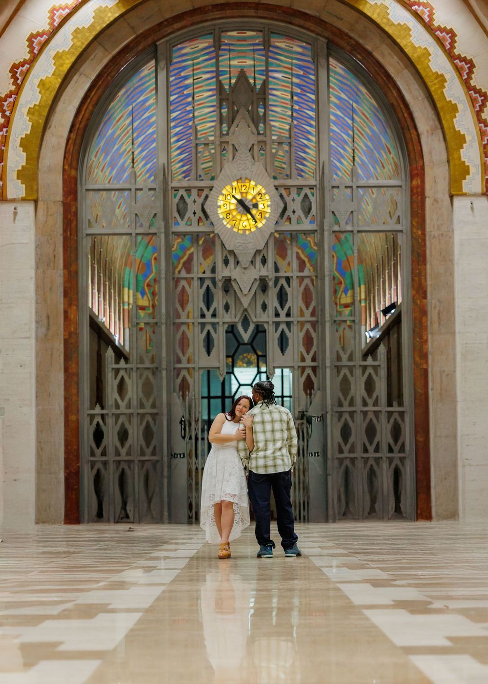 A couple stands silhouetted against an ornate Art Deco entrance with a grand clock and geometric metalwork patterns.