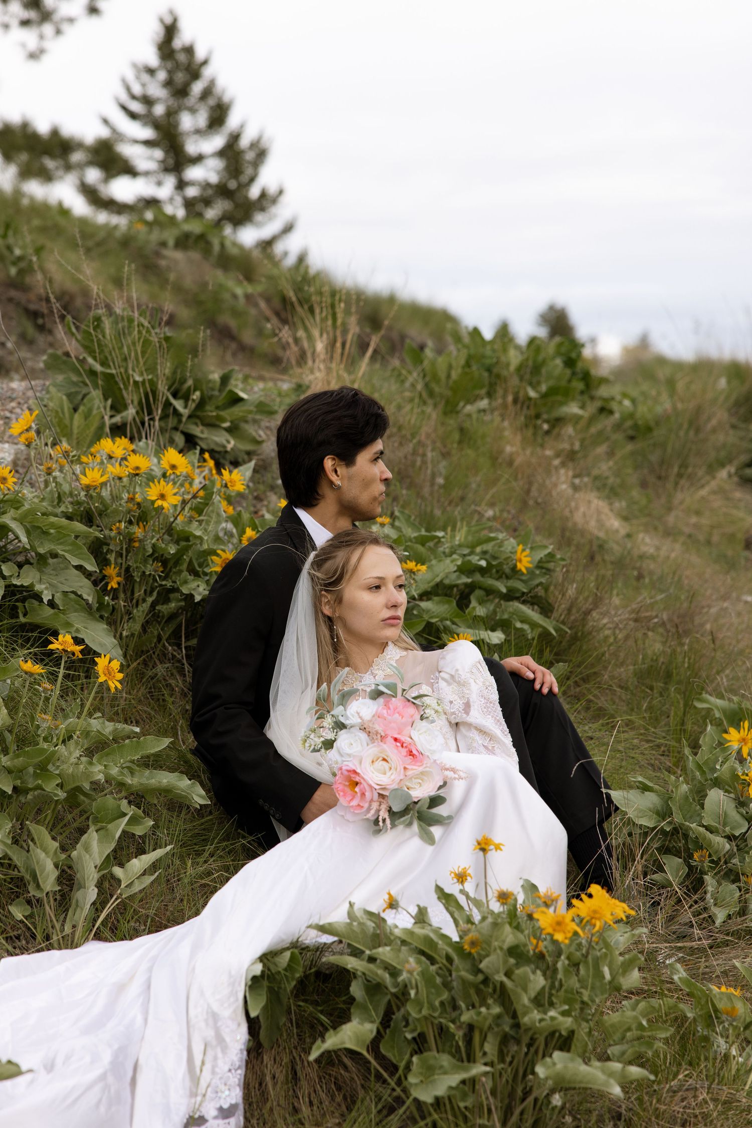 A couple in wedding attire sits among yellow wildflowers on a grassy hillside during an outdoor photo session.