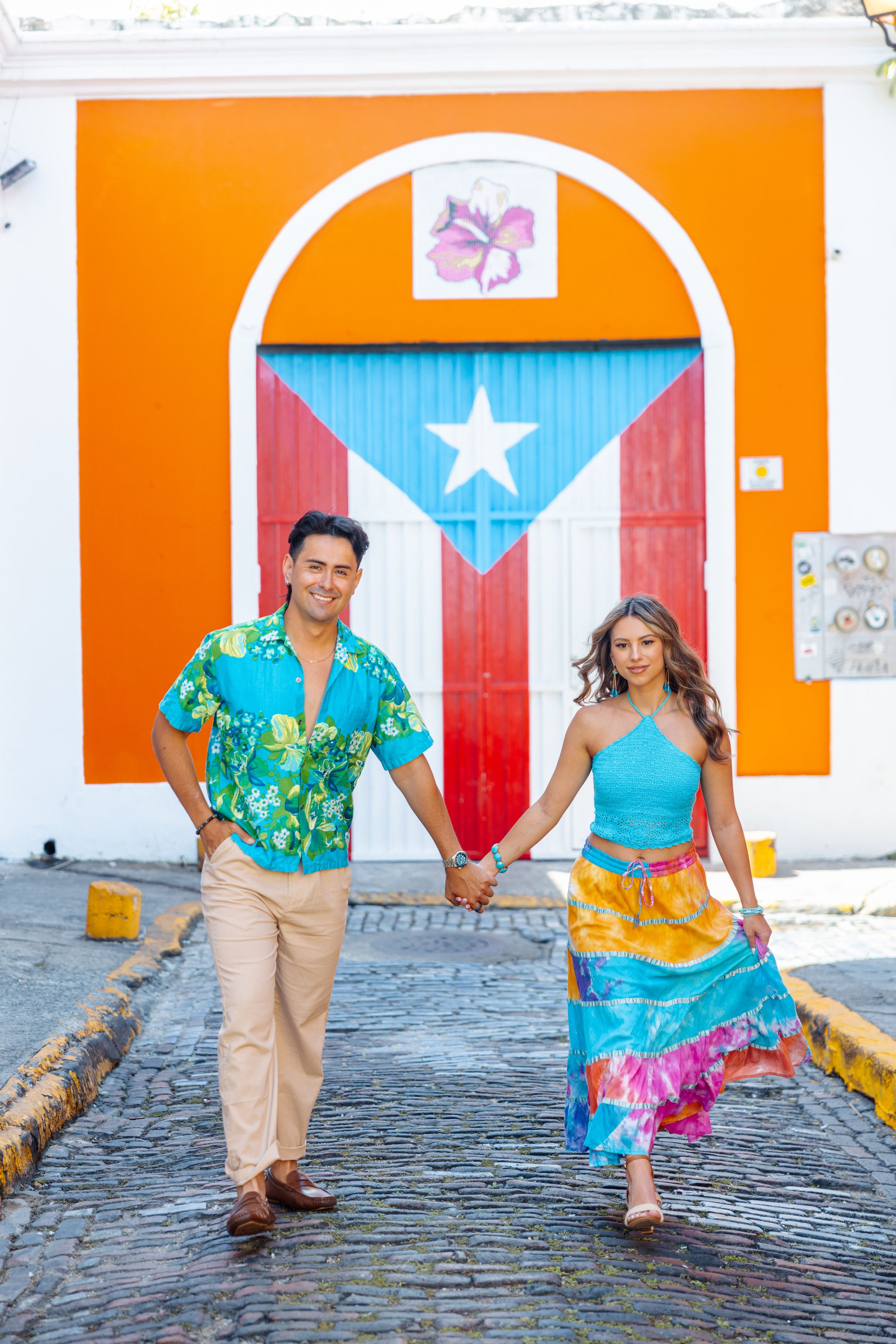 A vibrant couple holds hands walking in front of a colorful Puerto Rican flag mural on a cobblestone street.