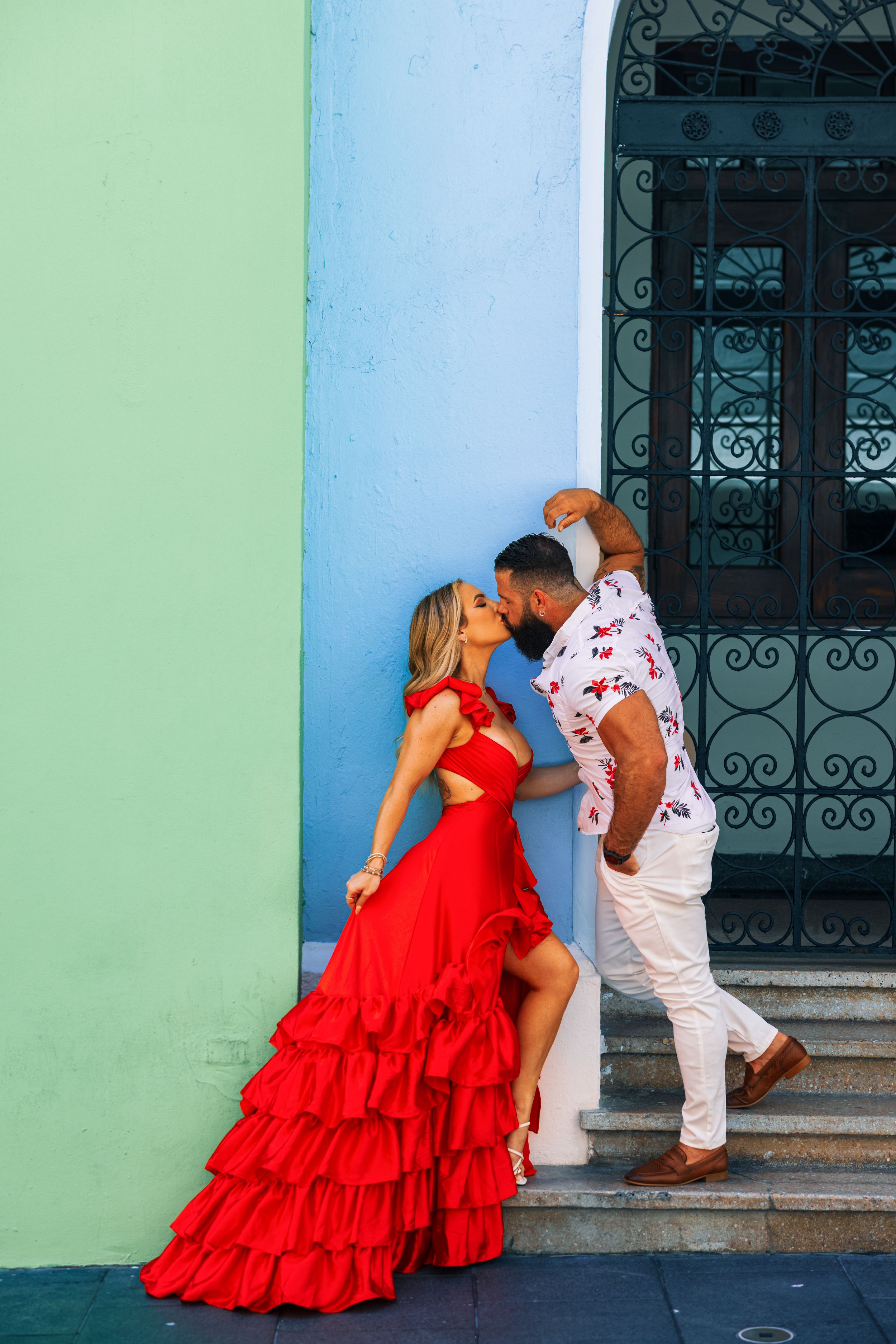 Couple sharing a passionate kiss against a mint green wall by ornate iron doors.