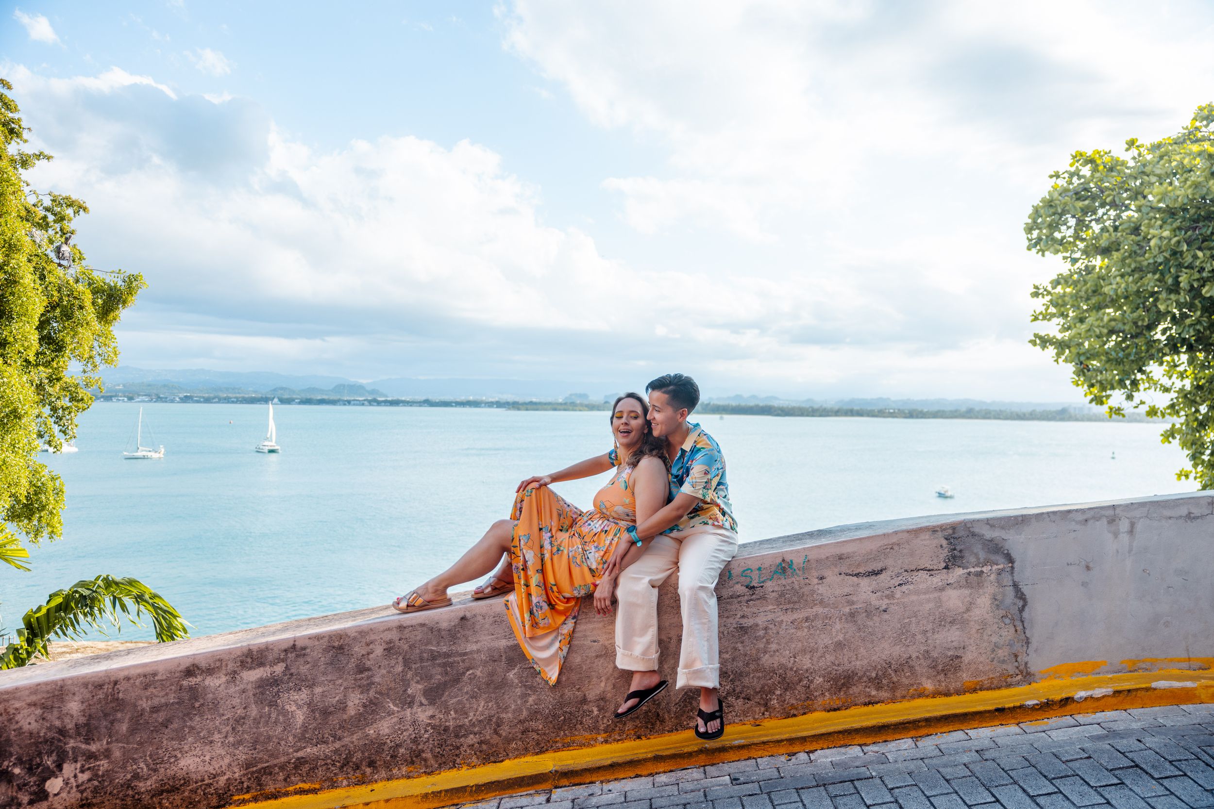 Romantic couple sitting on a stone wall overlooking turquoise Caribbean waters in Puerto Rico.
