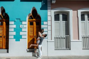 Joyful couple sharing a lift embrace in front of a teal and pink colonial facade.