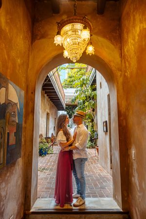 A couple embraces under a crystal chandelier in an arched colonial corridor with vibrant artwork on the walls.