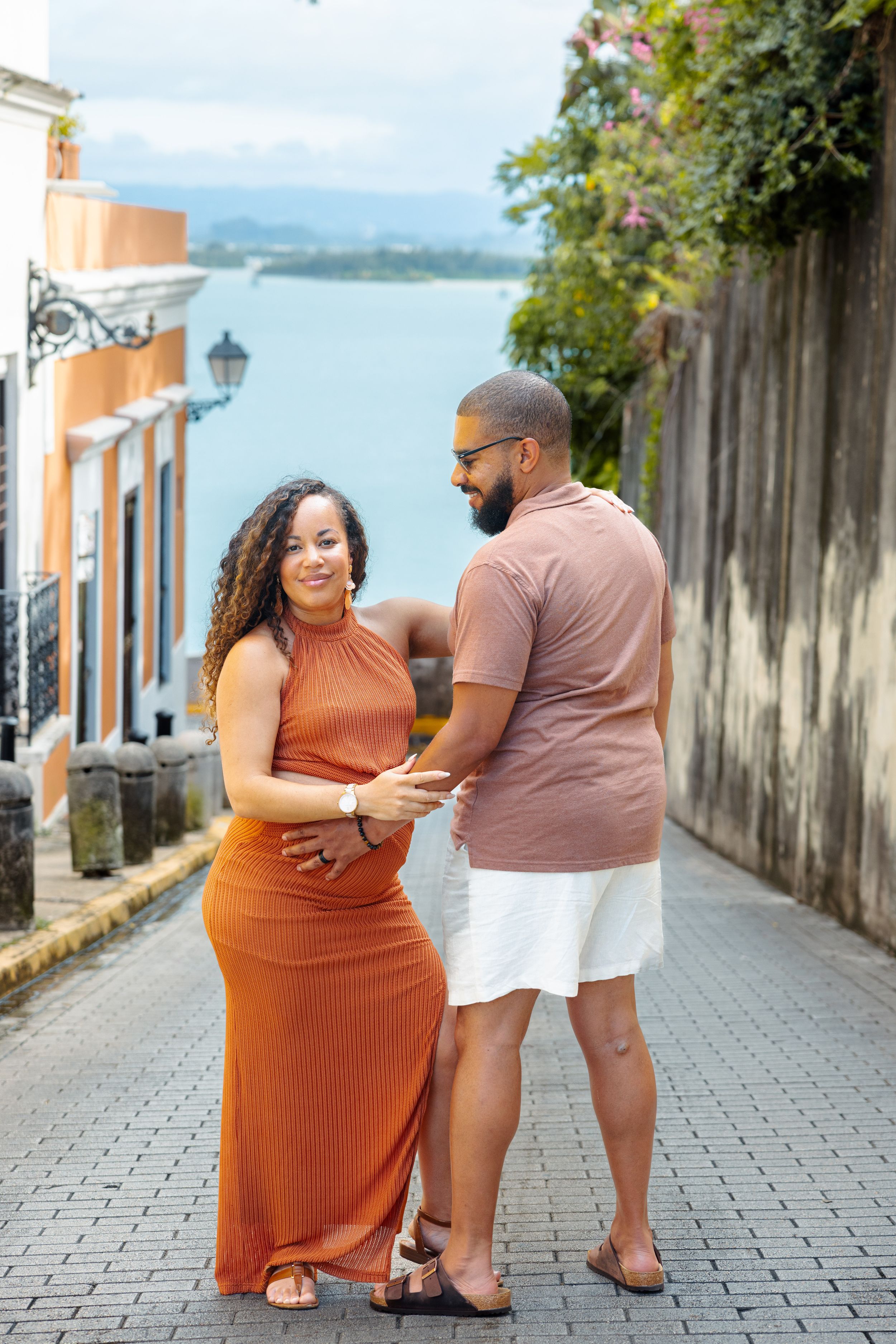 A pregnant woman in an orange dress and her partner hold hands on a scenic cobblestone street in Old San Juan.