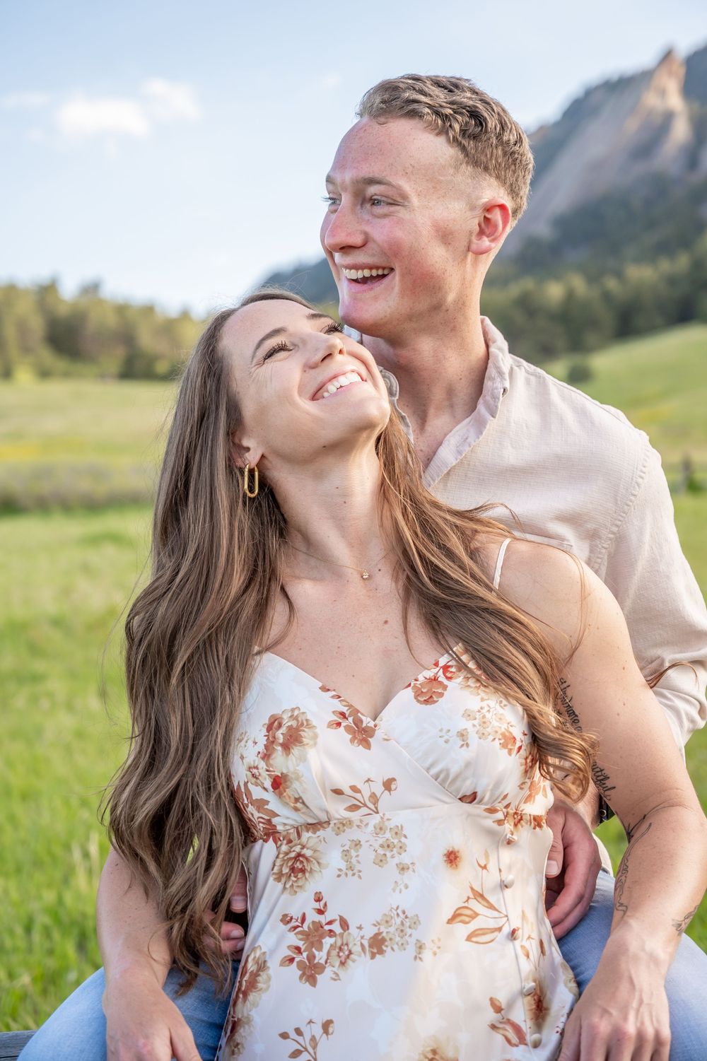 Happy couple laughs together in a meadow with mountains in background during golden hour.