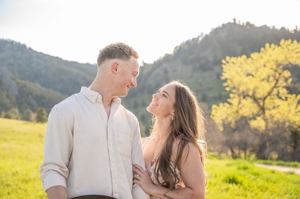A couple shares a tender moment in a sunny meadow with mountains and yellow blooming trees in the background.
