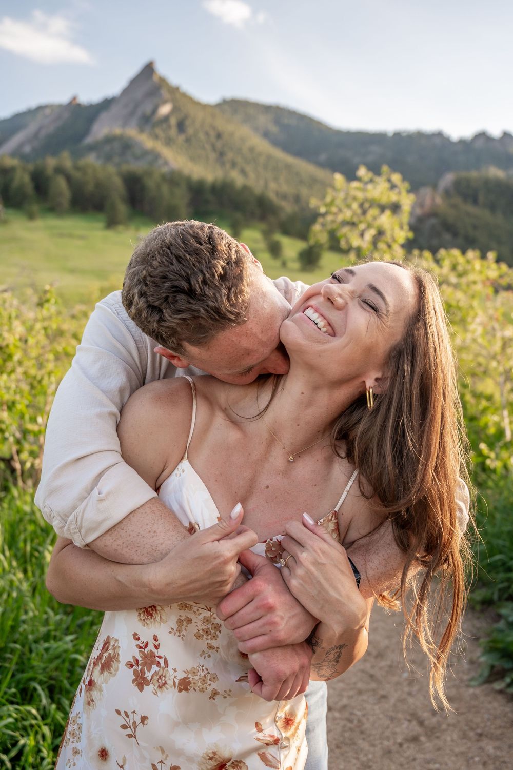 A romantic moment shared between a couple laughing and embracing on a mountain trail with scenic peaks in the background.
