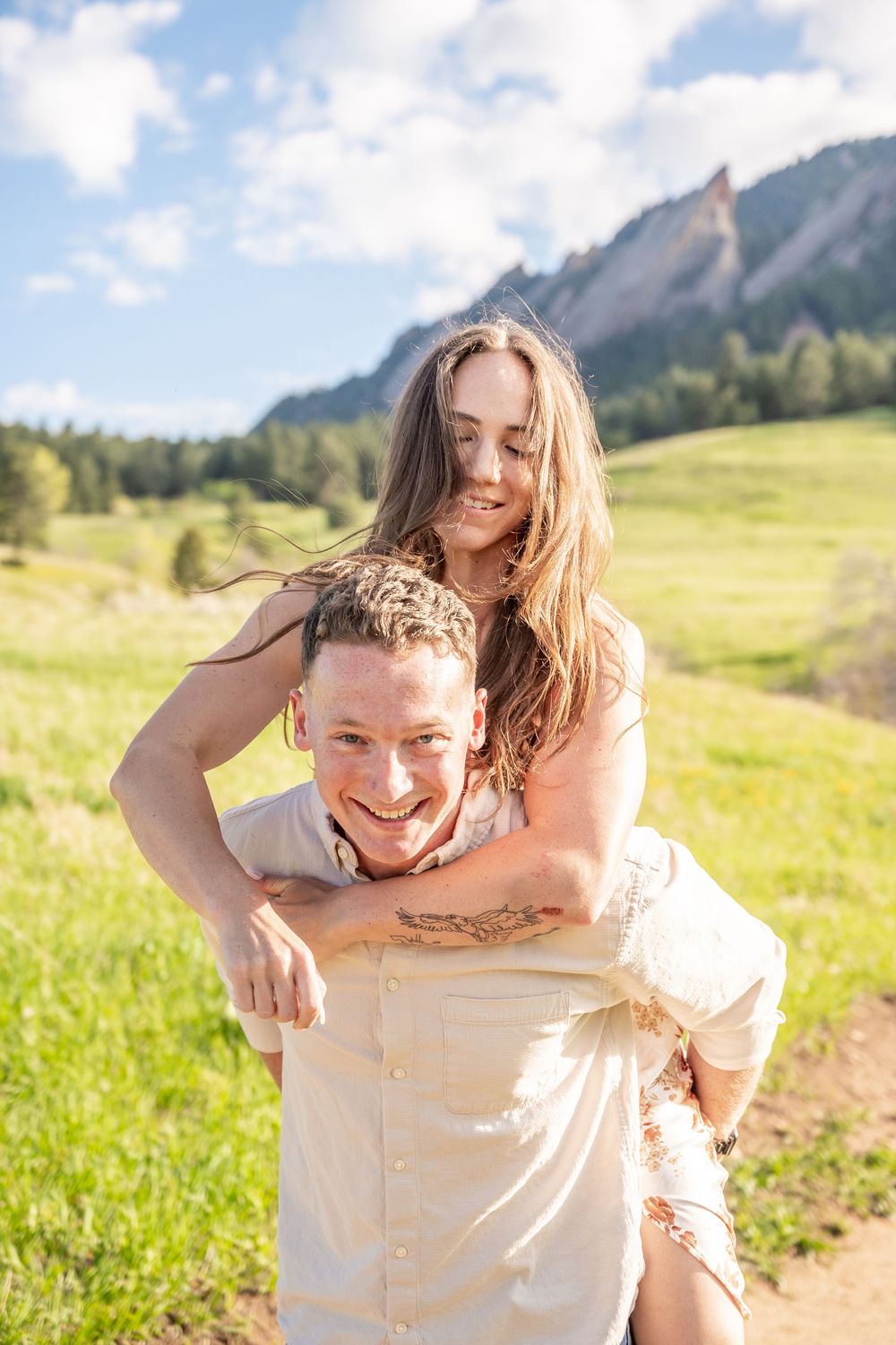 A couple enjoys a playful piggyback ride on a sunny day with the Flatirons mountains of Boulder, Colorado in the background.