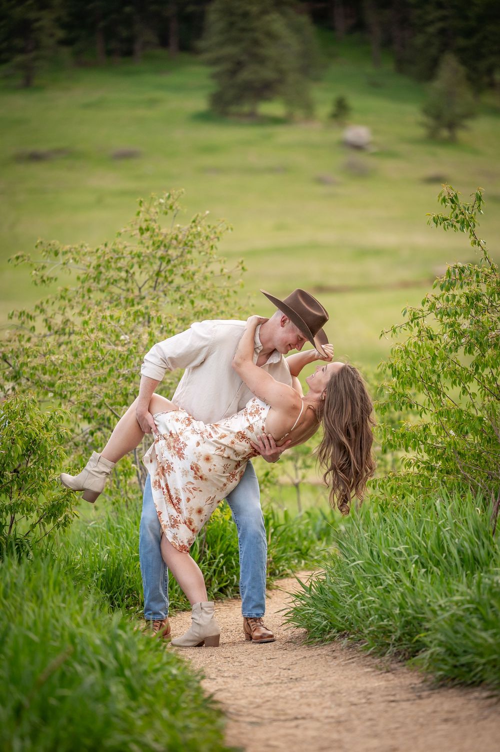 A romantic couple share an intimate moment on a dirt path surrounded by lush green foliage and rolling hills.
