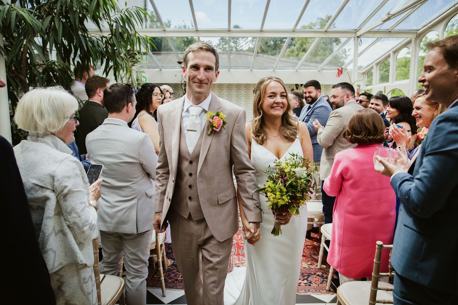 Joyful couple walks through greenhouse surrounded by wedding guests while bride holds yellow wildflower bouquet.