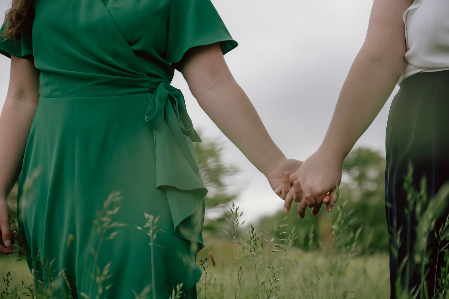 Two brides hold hands and stroll through a meadow on a spring morning, following their wedding together in Ledbury