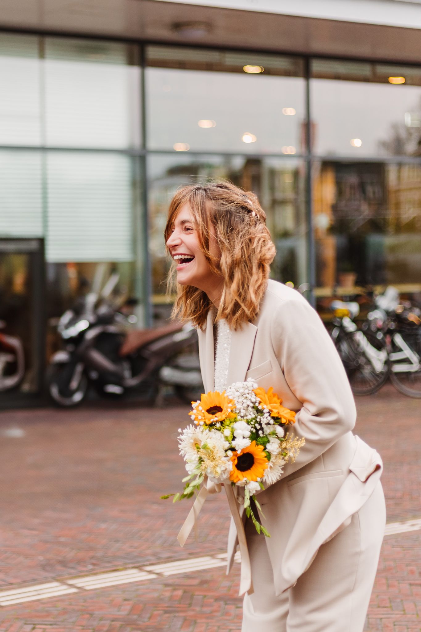 A bride in a cream-colored suit holds a vibrant bouquet of sunflowers while laughing outdoors after a gemeente wedding.