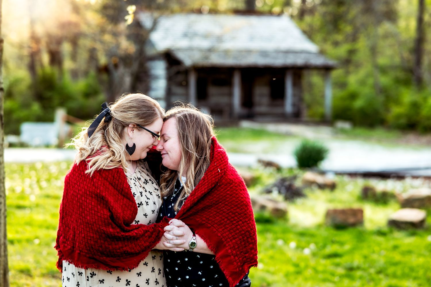 Two people wrapped in a cozy red blanket share an intimate moment in front of a rustic cabin on a sunny day.