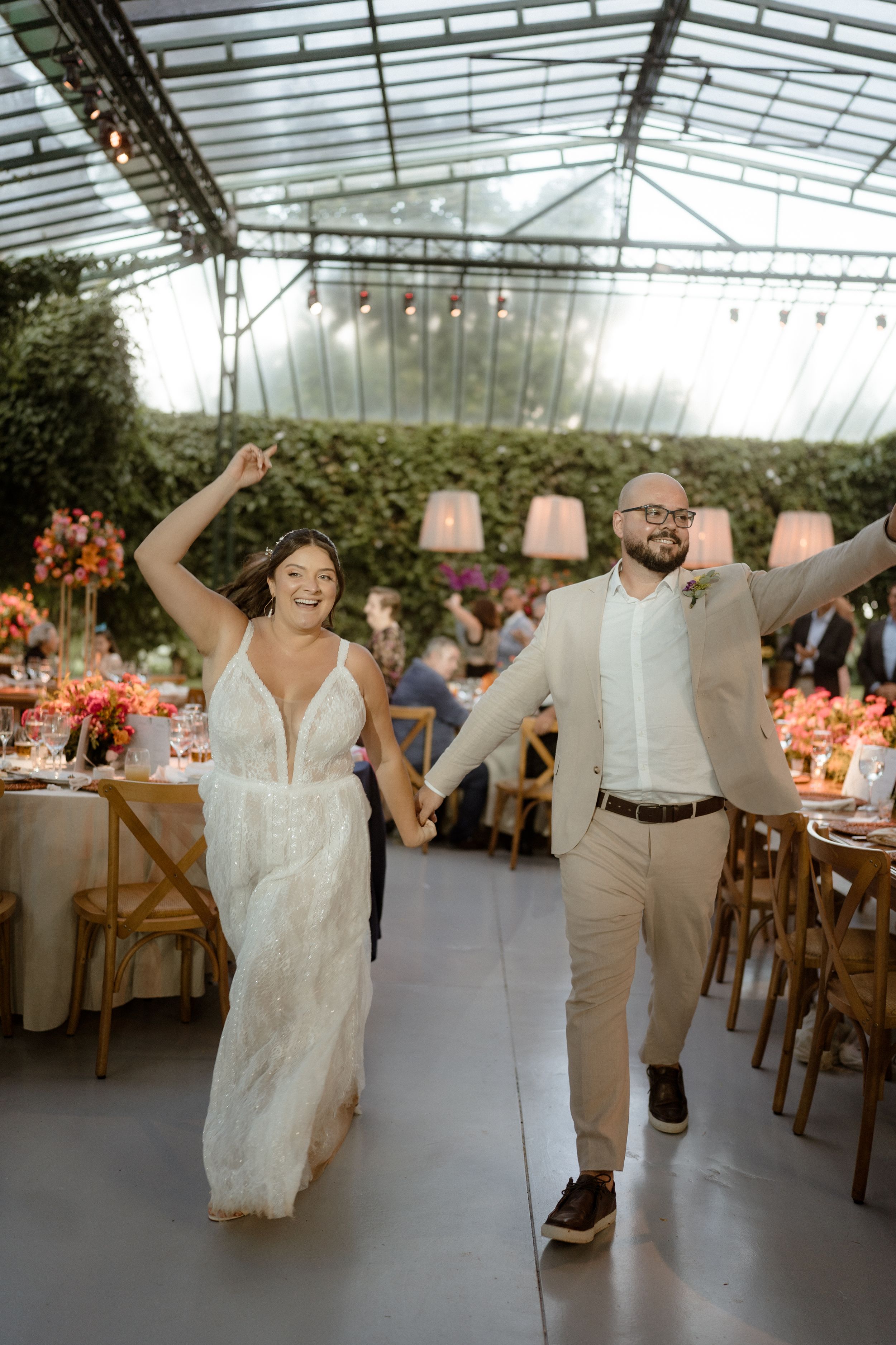 Wedding reception guests dancing and celebrating under a glass-roofed venue with warm lighting.