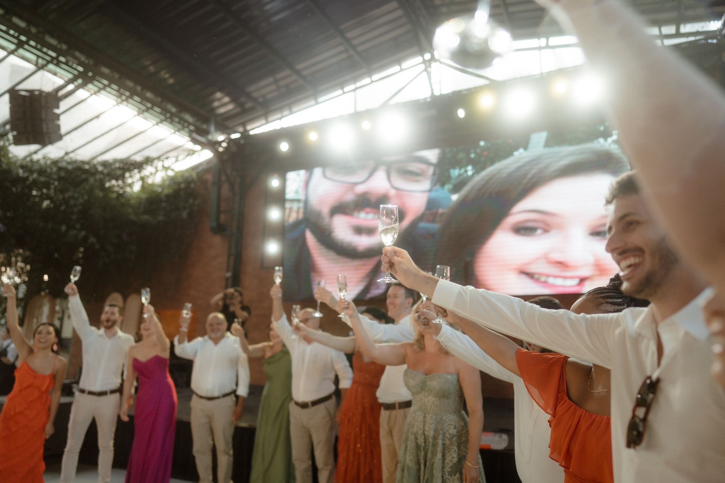 Wedding guests with raised arms dancing in celebration under an indoor greenhouse-style venue.