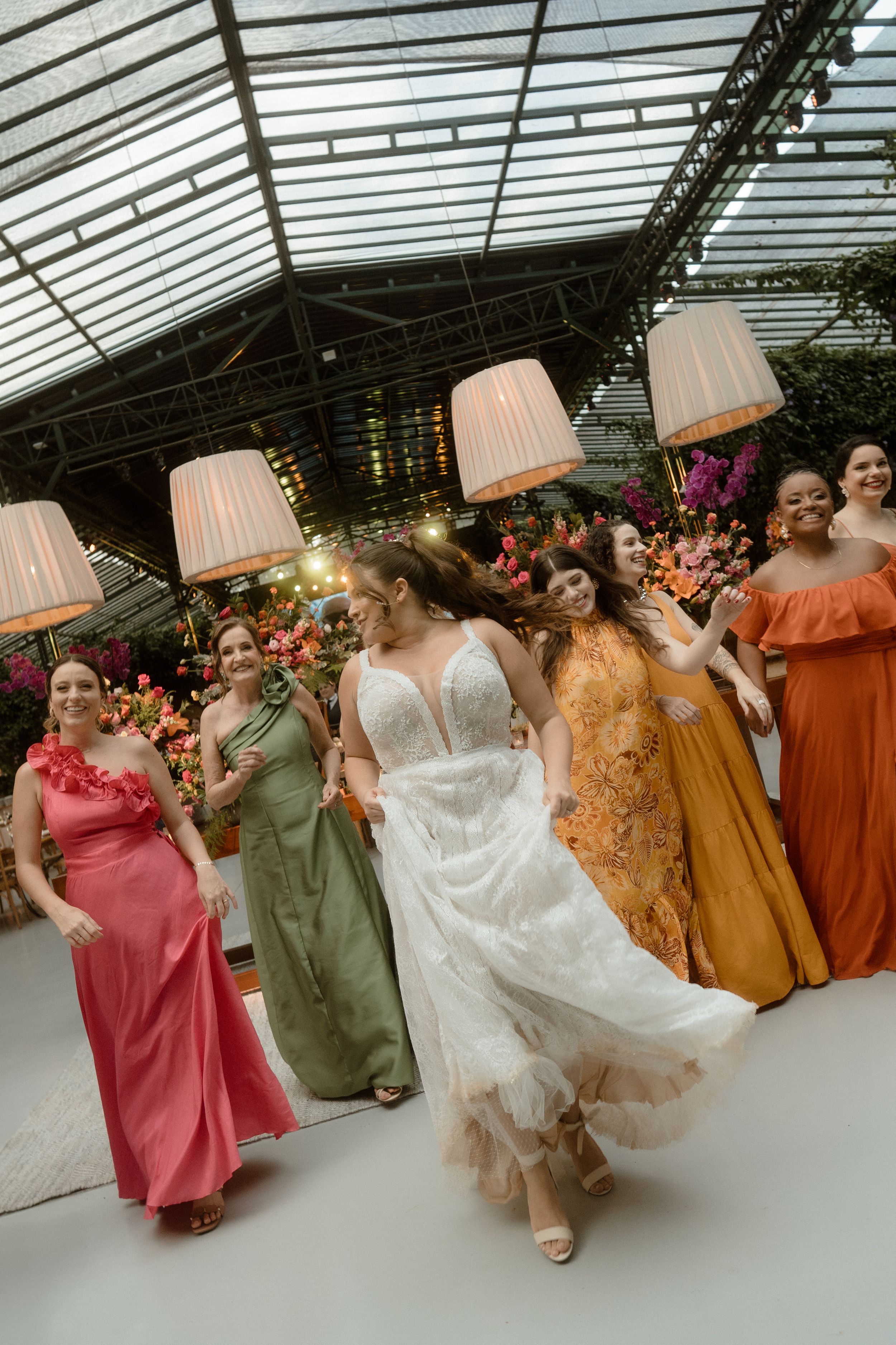 Wedding party in colorful dresses dancing in formation under a glass-roofed reception venue.