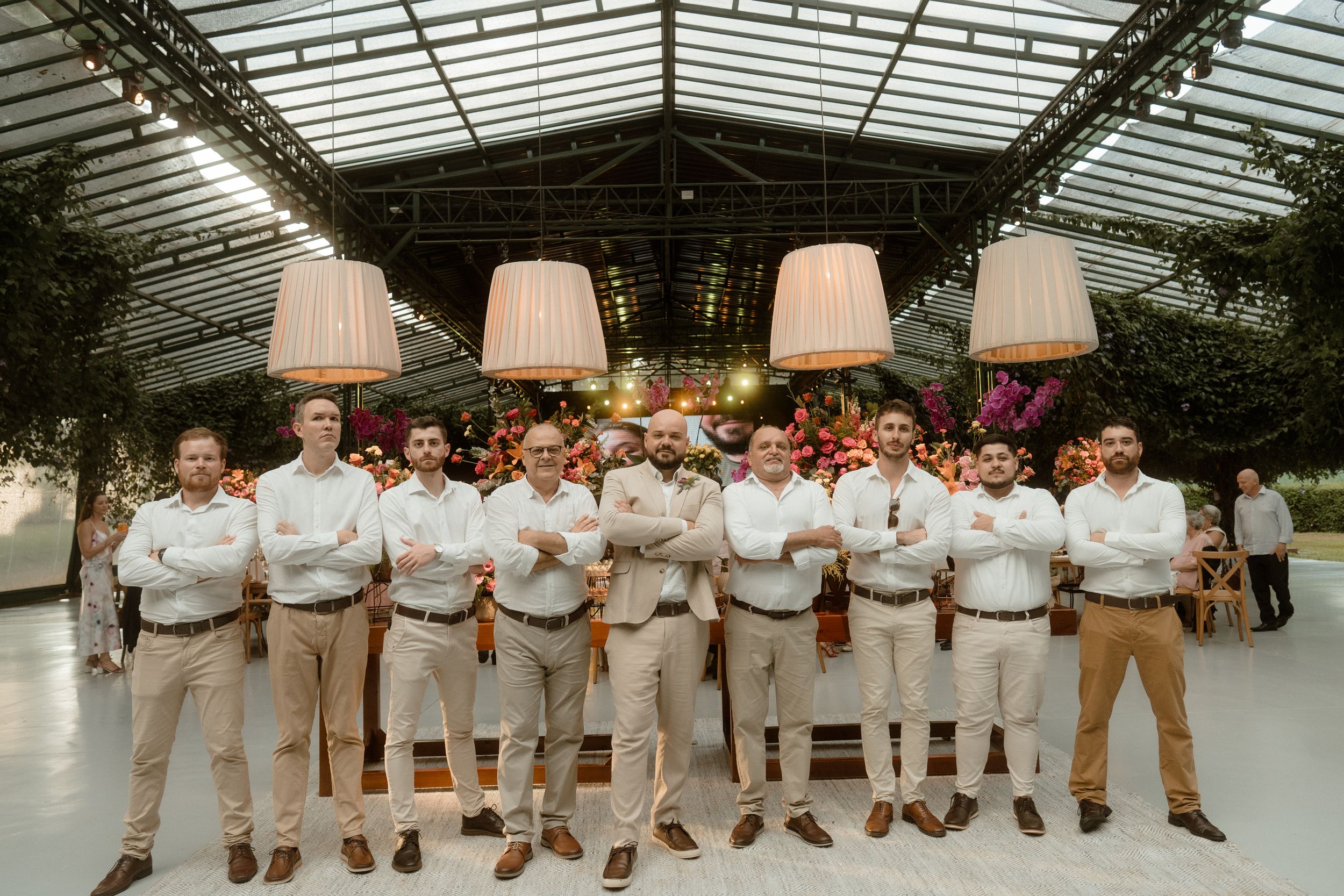 Groomsmen in matching tan suits pose in line under a glass-roofed venue.
