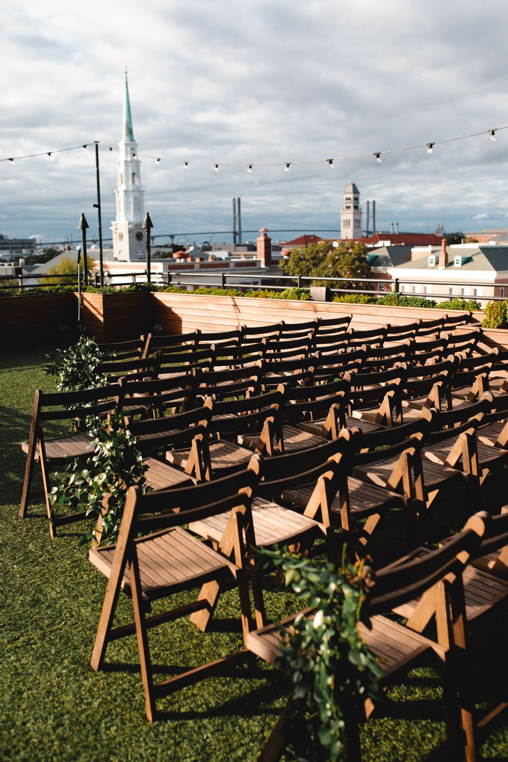 Rooftop wedding ceremony setup with wooden folding chairs, string lights, and city skyline views on a cloudy day.