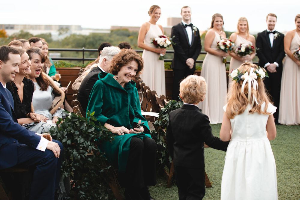 Woman in green velvet dress smiles at flower girl and ring bearer during outdoor wedding ceremony with bridesmaids.