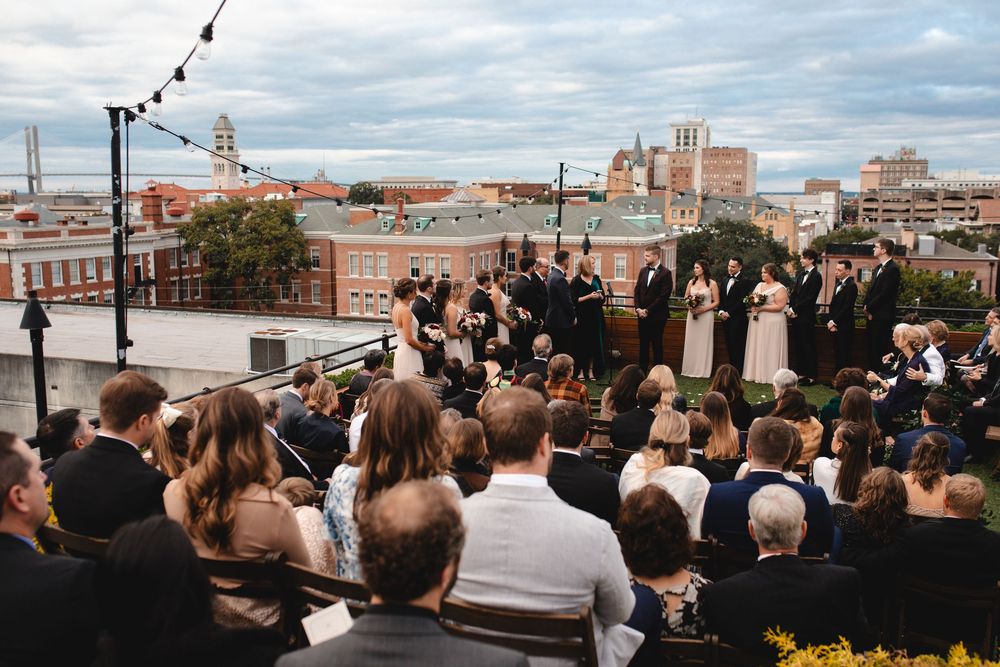 Rooftop wedding ceremony with city skyline backdrop, guests seated outdoors under string lights.