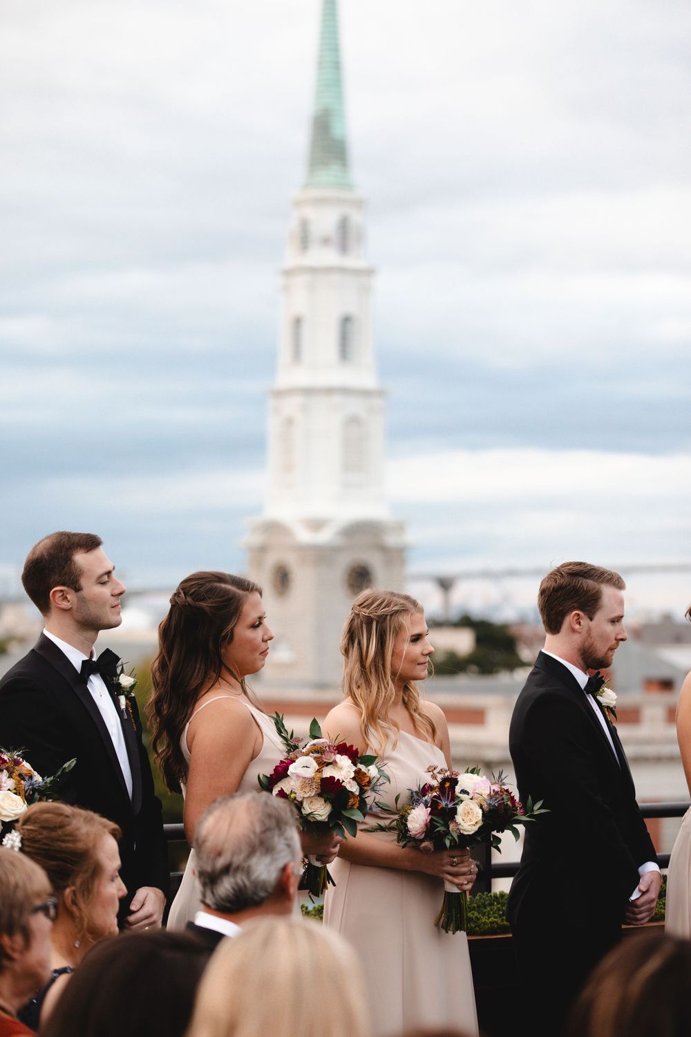 Wedding party stands outdoors with a white church steeple backdrop under cloudy skies, bridesmaids holding floral bouquets.