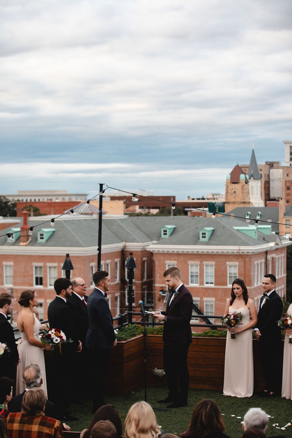 Rooftop wedding ceremony with couple exchanging vows, bridal party, and city skyline backdrop under cloudy skies.